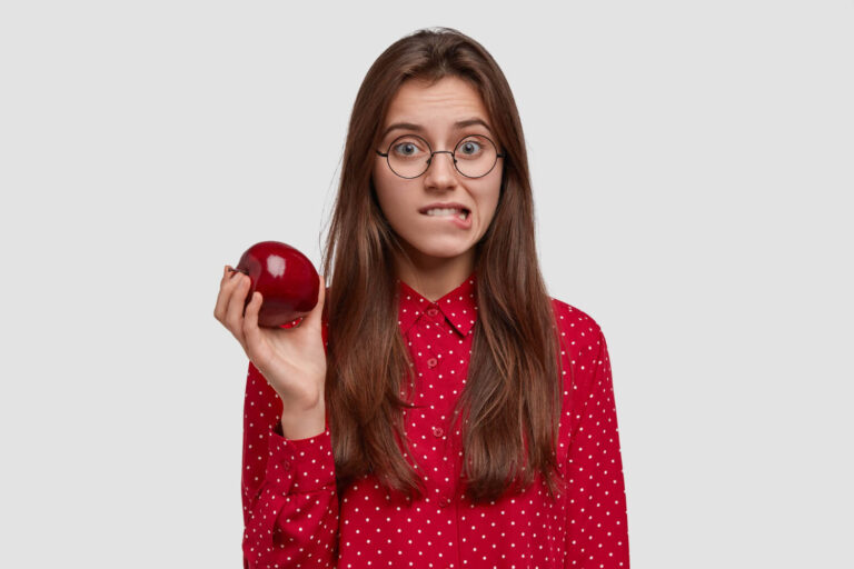 Mulher jovem de óculos e camisa vermelha, expressando dúvida ao segurar uma maçã, ilustrando cuidados com lentes de contato dental na Aoun Odontologia em Guarulhos.