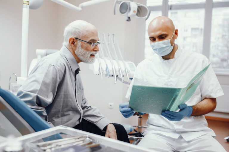 Dentista com máscara e luvas azuis explica ao paciente idoso, de barba branca e óculos, os detalhes de um plano de tratamento para prótese dentária em Guarulhos. O paciente olha atentamente para a pasta aberta.